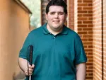 Smiling man in teal polo shirt standing near brick wall holding a white cane.