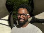 Person with glasses and beard in gray shirt smiling outdoors with abstract sculpture in background.