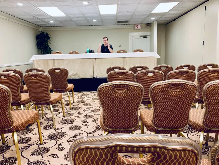 guy sitting on a stage presenting for an empty room with chairs.