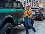 Person in mustard jacket and jeans standing beside a vintage green off-road vehicle on brick-paved street in urban setting.